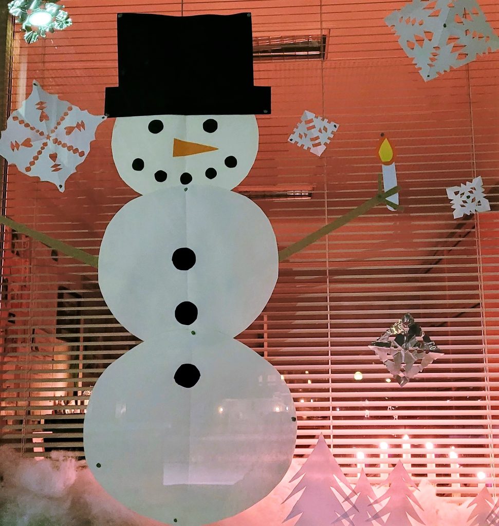 Koulun ikkunan täyttää iso, valkoinen lumiukko, jolla on porkkananenä ja musta hattu päässä. Ympärillä sataa lumihiutaleita. Tehty paperista. Iloinen ilme.